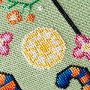 May include: Close-up of a cross-stitch design on a light green fabric. The design features orange and white flowers, a yellow and white circular pattern, and a blue and orange candy cane shape. Black border.