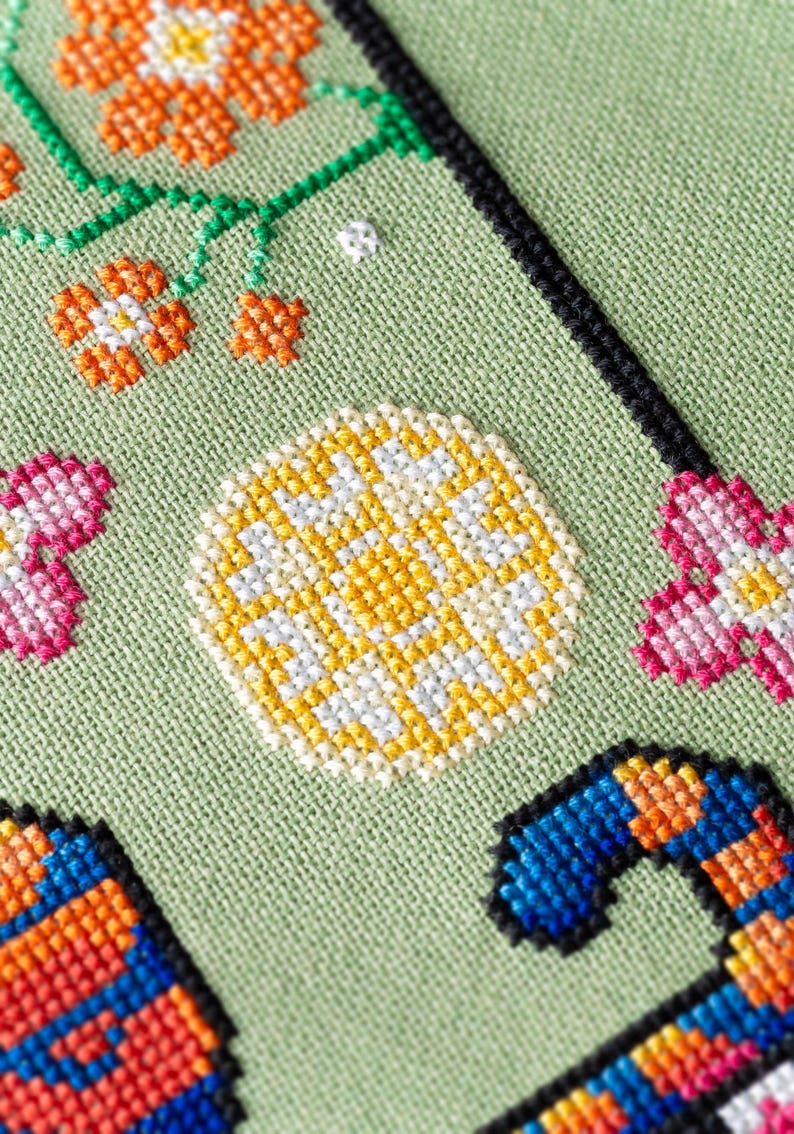 May include: Close-up of a cross-stitch design on a light green fabric. The design features orange and white flowers, a yellow and white circular pattern, and a blue and orange candy cane shape. Black border.
