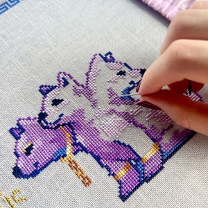 May include: Cross-stitch embroidery in progress, featuring a design of three purple and white mythical creatures. The needle is threaded with blue thread, and the fabric is stretched within a pink hoop. The design includes gold and blue accents.