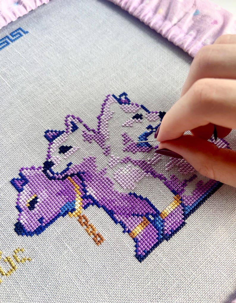 May include: Cross-stitch embroidery in progress, featuring a design of three purple and white mythical creatures. The needle is threaded with blue thread, and the fabric is stretched within a pink hoop. The design includes gold and blue accents.