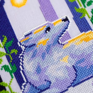 May include: A cross-stitch design featuring a wolf howling at the moon. The artwork uses shades of blue, purple, and yellow. The wolf is the focal point, with a detailed depiction of its fur and expression. The background includes stylized trees and foliage.