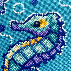 May include: A close-up of a cross-stitch design featuring a seahorse in shades of teal, purple, and yellow against a turquoise background. White stitches form bubbles and other aquatic shapes, creating a textured, handcrafted appearance.