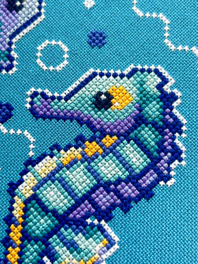 May include: A close-up of a cross-stitch design featuring a seahorse in shades of teal, purple, and yellow against a turquoise background. White stitches form bubbles and other aquatic shapes, creating a textured, handcrafted appearance.