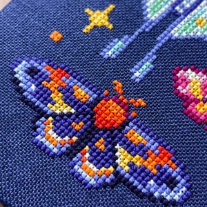 Op de afbeelding: Close-up van een kruissteekontwerp op marineblauwe stof. Het ontwerp toont een vlinder met oranje, gele en blauwe details. Extra elementen zijn gele sterren en een roze en geel hart.