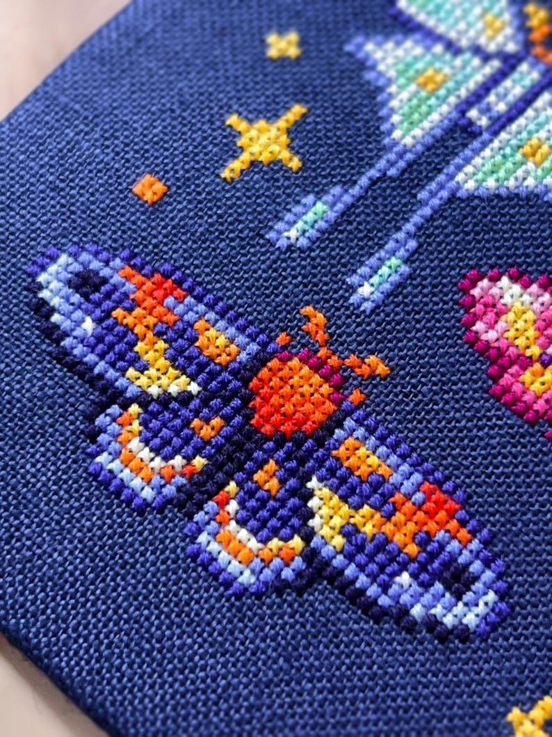 Op de afbeelding: Close-up van een kruissteekontwerp op marineblauwe stof. Het ontwerp toont een vlinder met oranje, gele en blauwe details. Extra elementen zijn gele sterren en een roze en geel hart.