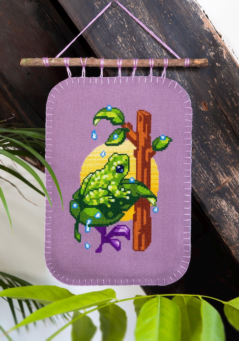 Peut inclure: Une tenture murale rectangulaire en tissu violet avec un motif au point de croix repr&eacute;sentant une grenouille verte sur une branche brune devant un cercle jaune. L'ensemble est suspendu &agrave; une brindille en bois par des boucles en fil violet.