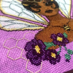 Closeup view of the lefthand bottom side. Here there are purple flowers with yellow cores and a yellow honeycomb pattern consisting of backstitch. The honeycomb is subtle. The purple flowers have green stems.