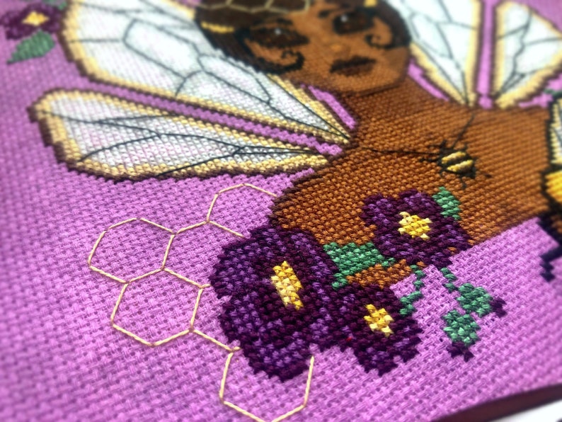 Closeup view of the lefthand bottom side. Here there are purple flowers with yellow cores and a yellow honeycomb pattern consisting of backstitch. The honeycomb is subtle. The purple flowers have green stems.