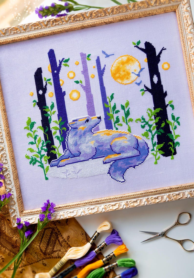 May include: Cross-stitch artwork in a gold frame depicts a wolf in a forest scene. The wolf is blue and yellow, lying down in front of trees, a full moon, and birds. The artwork is on white fabric. The frame is ornate.