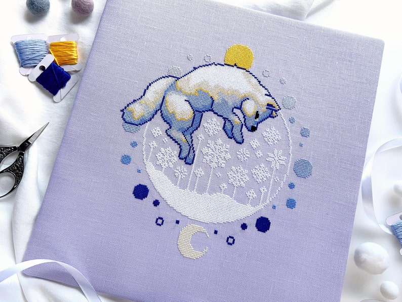 May include: A cross-stitch pattern on a purple fabric featuring a white fox jumping in a circle with a yellow sun, blue snowflakes, and a crescent moon.