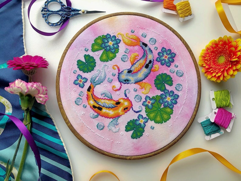 May include: A pink and white embroidery hoop with a cross-stitch design of two koi fish swimming in a pond with lily pads and bubbles. The fish are orange and blue with black markings.