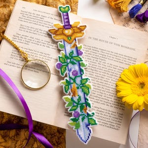 May include: A handmade, cross-stitched bookmark in the shape of a sword, featuring a detailed design with gold, green, purple, and white colors. The bookmark is placed on an open book with text, alongside embroidery floss and a yellow flower.