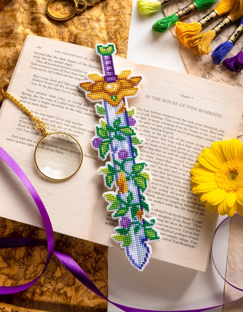 K&ouml;nnte beinhalten: Handgefertigtes Lesezeichen in Schwertform, kreuzgestickt mit goldenen, gr&uuml;nen, lila und wei&szlig;en Details. Es liegt auf einem offenen Buch, zusammen mit Stickgarn und einer gelben Blume. Die L&auml;nge betr&auml;gt ca. 20 cm.
