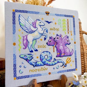 May include: Cross-stitch art featuring mythological creatures: a winged horse, a three-headed dog, and a sea serpent. The design includes Greek text and a blue and gold border. The artwork is displayed on a wooden easel.