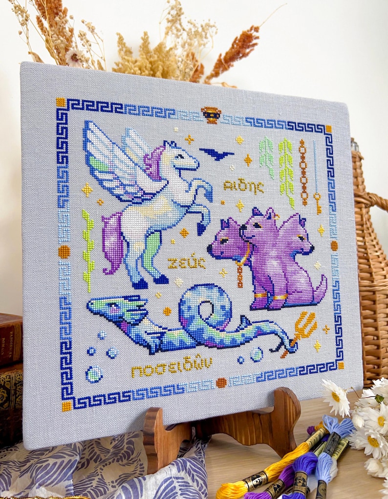 May include: Cross-stitch art featuring mythological creatures: a winged horse, a three-headed dog, and a sea serpent. The design includes Greek text and a blue and gold border. The artwork is displayed on a wooden easel.