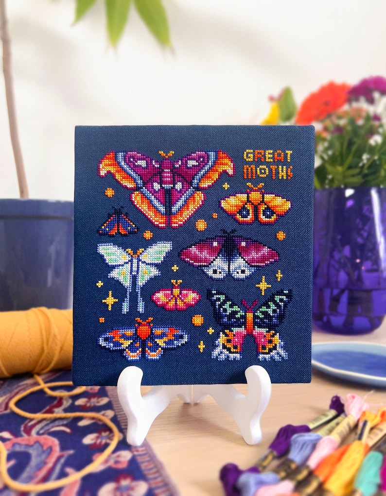 Op de afbeelding: Een kruissteekkunstwerk met diverse kleurrijke motontwerpen op een marineblauwe achtergrond. De tekst "GREAT MOTHS" is oranje geborduurd. Het kunstwerk wordt gepresenteerd op een witte ezel.