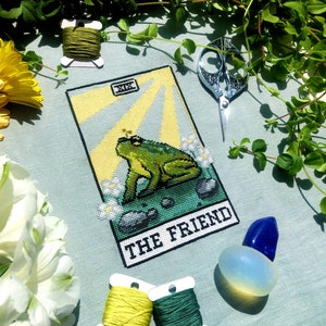 Flatlay of The Friend frog tarot cross stitch pattern. Stitched item, surrounded by decorations. Finished piece is of small to medium size. Colors are yellow, green, blue, grey and black. Frog is cute and small. The Friend is written underneath.