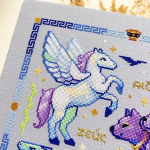 May include: Cross-stitch embroidery featuring a winged horse, a purple and white horse, and other mythological figures. The design includes the word "Zeus" in gold thread, a blue border, and a light blue background.