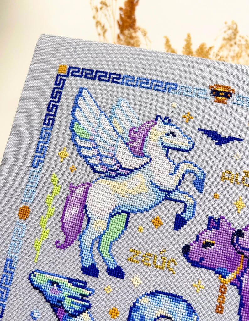 May include: Cross-stitch embroidery featuring a winged horse, a purple and white horse, and other mythological figures. The design includes the word "Zeus" in gold thread, a blue border, and a light blue background.