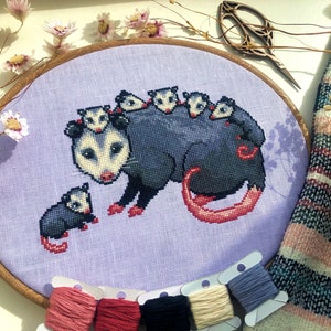 Front view of possum mama cross stitch pattern. The opossum mother has 6 babies, 5 of which are on her back. The 6th one is sitting on her left. Colors are mainly blueish grey, lilac, beige and pink. The baby possums are extremely cute.