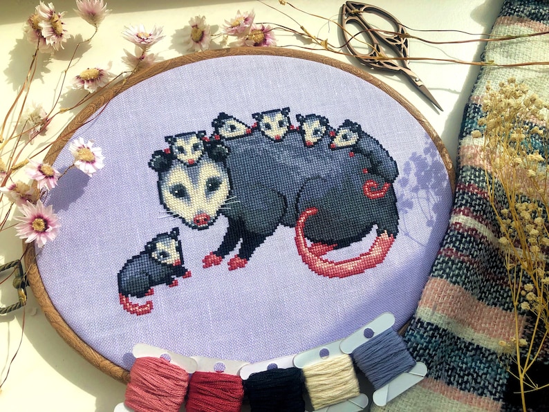 Front view of possum mama cross stitch pattern. The opossum mother has 6 babies, 5 of which are on her back. The 6th one is sitting on her left. Colors are mainly blueish grey, lilac, beige and pink. The baby possums are extremely cute.