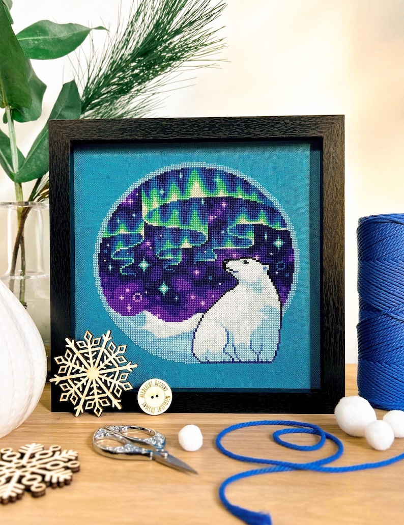 May include: A framed cross-stitch design featuring a polar bear standing in front of a blue and purple aurora borealis. The design is surrounded by a white circle and a blue background.