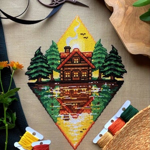 Flat-lay of Lakeside Cabin cross stitch pattern. Stitched item, surrounded by decorations. Finished piece is of large size. Colors are yellow, brown, orange, green, black and white. Scenery contains a sunset, and the cabin is reflected in the water.