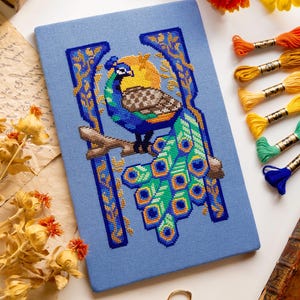 May include: A rectangular light blue fabric panel featuring a cross-stitch design of a peacock perched on a branch. The peacock has a blue head, a brown patterned wing, and a long green tail with blue and yellow eye-shaped feathers.