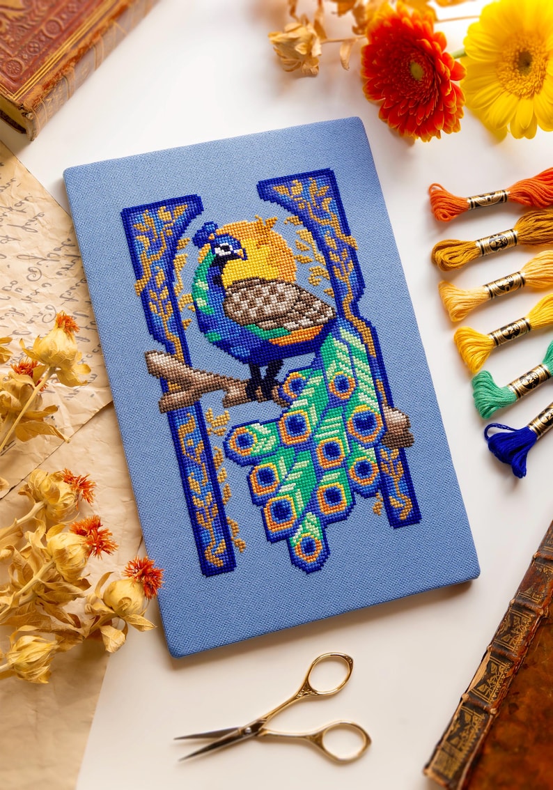 May include: A rectangular light blue fabric panel featuring a cross-stitch design of a peacock perched on a branch. The peacock has a blue head, a brown patterned wing, and a long green tail with blue and yellow eye-shaped feathers.