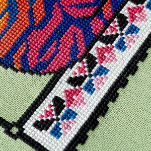 May include: Close-up of a cross-stitch design with vibrant colors. The design features orange, pink, and blue geometric patterns on a light green fabric. A border of black cross-stitches frames the design.
