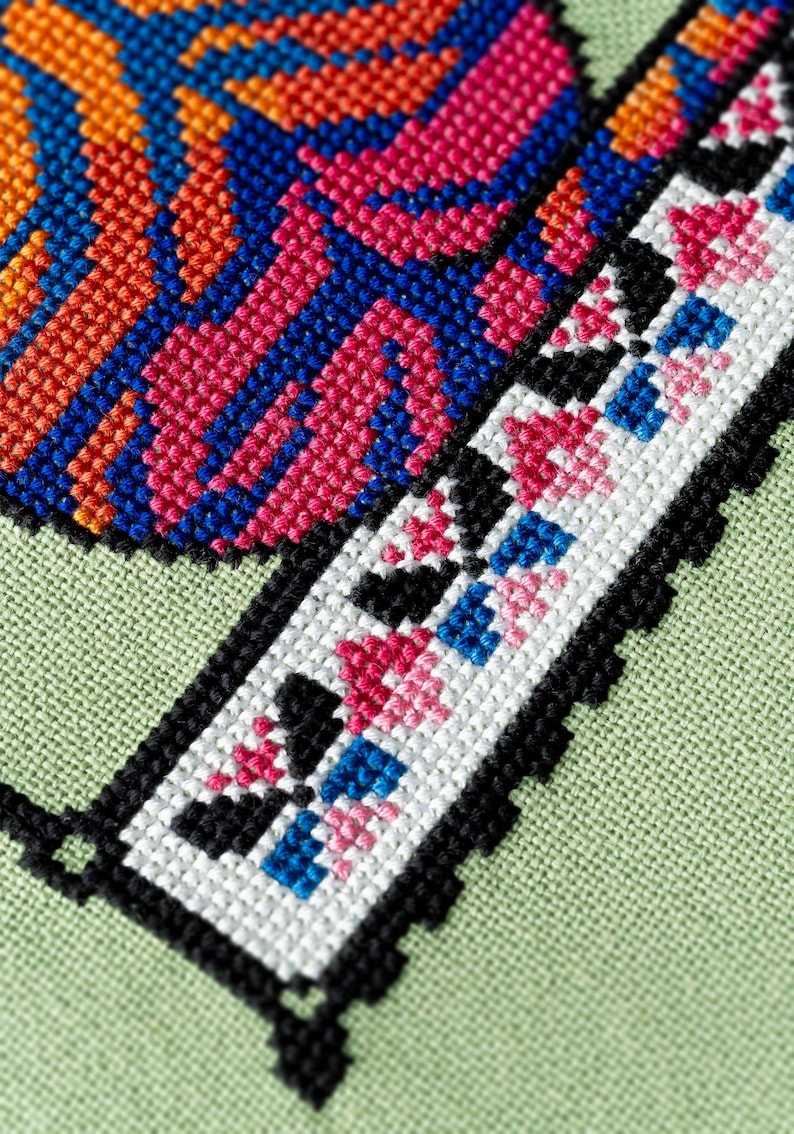 May include: Close-up of a cross-stitch design with vibrant colors. The design features orange, pink, and blue geometric patterns on a light green fabric. A border of black cross-stitches frames the design.