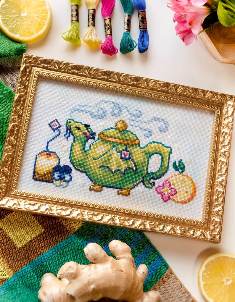 May include: A cross-stitch embroidery in a gold ornate frame featuring a green dragon-shaped teapot with a tea bag, steam swirls, and a citrus slice. The design is stitched on white fabric with colorful thread accents and clear detail.