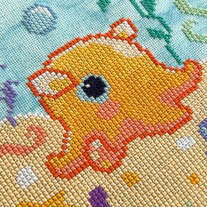 May include: A cross-stitch pattern of a yellow and orange fish with a black eye, surrounded by blue and green seaweed and white bubbles. The background is a light blue color.