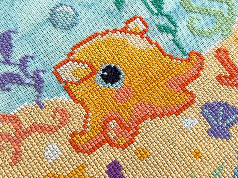 May include: A cross-stitch pattern of a yellow and orange fish with a black eye, surrounded by blue and green seaweed and white bubbles. The background is a light blue color.