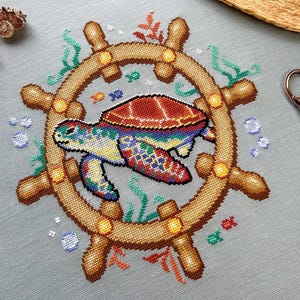 May include: A colorful cross-stitch design of a sea turtle swimming inside a wooden ship's wheel. The turtle is surrounded by green seaweed and blue bubbles.