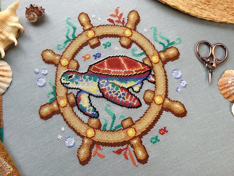 May include: A colorful cross-stitch design of a sea turtle swimming inside a wooden ship's wheel. The turtle is surrounded by green seaweed and blue bubbles.
