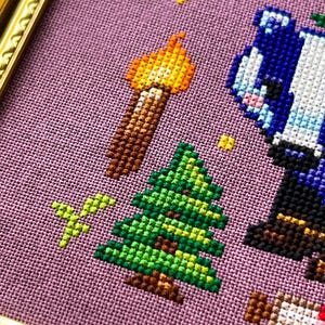 Puede incluir: Arte de punto de cruz que presenta un &aacute;rbol de Navidad, una antorcha y un personaje azul sobre un fondo p&uacute;rpura. La obra de arte est&aacute; enmarcada en un marco dorado. El dise&ntilde;o de punto de cruz utiliza varios colores, incluyendo verde, marr&oacute;n, amarillo y azul.