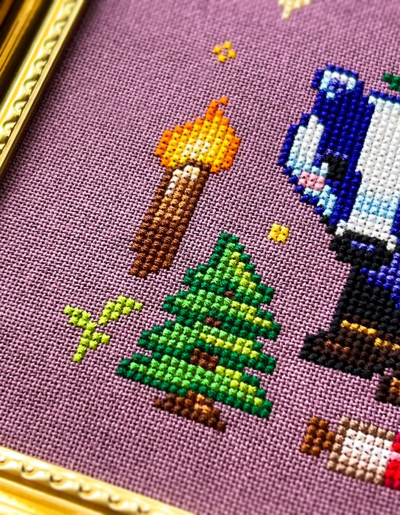 Puede incluir: Arte de punto de cruz que presenta un &aacute;rbol de Navidad, una antorcha y un personaje azul sobre un fondo p&uacute;rpura. La obra de arte est&aacute; enmarcada en un marco dorado. El dise&ntilde;o de punto de cruz utiliza varios colores, incluyendo verde, marr&oacute;n, amarillo y azul.