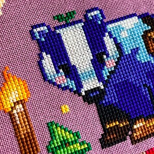 Puede incluir: Arte de punto de cruz con un personaje pixelado azul y blanco con botas marrones, un &aacute;rbol verde y una antorcha. El fondo es una tela de color p&uacute;rpura claro. Otros elementos pixelados incluyen un sol amarillo y un objeto a rayas rojas y blancas.
