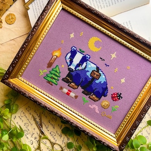 Puede incluir: Arte de punto de cruz en un marco decorativo. La obra presenta un tej&oacute;n azul con una mochila, una luna creciente, estrellas y una antorcha. El marco tiene un acabado dorado y marr&oacute;n, y el fondo es de tela morada clara.