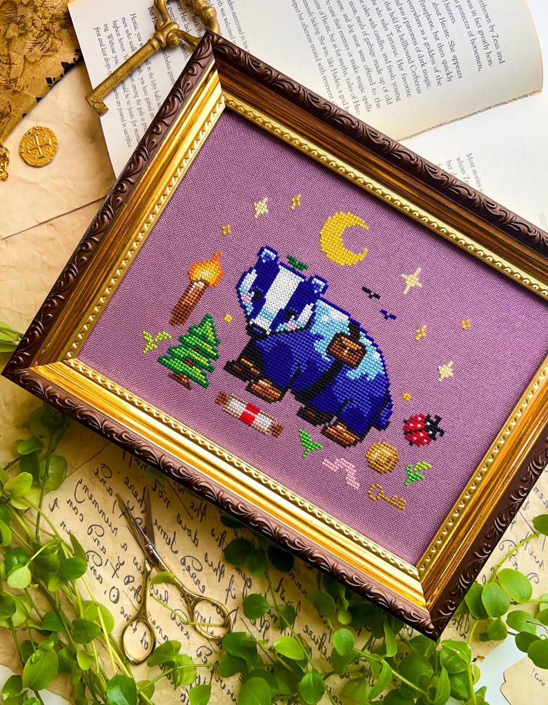 Puede incluir: Arte de punto de cruz en un marco decorativo. La obra presenta un tej&oacute;n azul con una mochila, una luna creciente, estrellas y una antorcha. El marco tiene un acabado dorado y marr&oacute;n, y el fondo es de tela morada clara.
