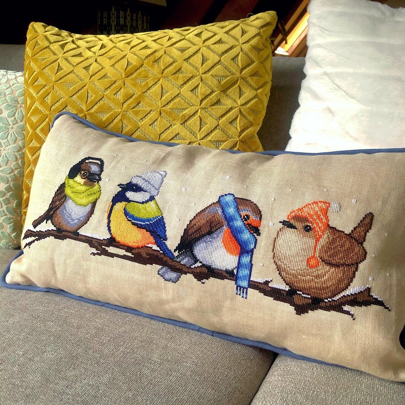 May include: A rectangular decorative pillow with a cross-stitched design of four birds perched on a branch. The birds are wearing scarves and hats in various colors. The pillow has a light beige background with a blue border.