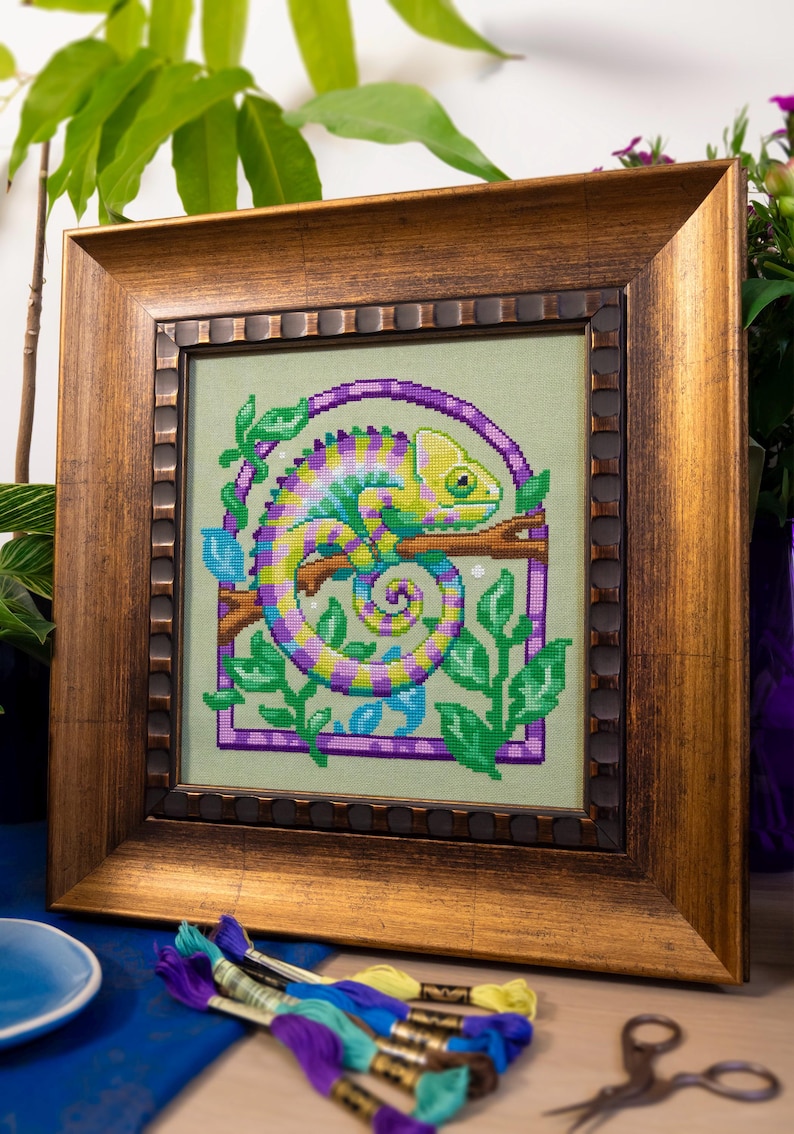 May include: Framed cross-stitch art featuring a colorful chameleon on a branch, surrounded by green leaves and a purple frame. The artwork is set against a light green background and framed in a gold-toned wooden frame. Embroidery floss and scissors are in the foreground.