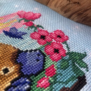 Extreme closeup of righthand side of platypus cross stitch pattern. Platypus is sniffing the pink flowers, which have black cores. Stitches are neat and tidy. Colors are vibrant. Platypus is adorable.