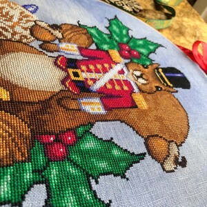 Closeup of squirrel cross stitch pattern, bottom righthand side. Stitches are neat and tidy. The pattern is rather large. Colors are vibrant and saturated. Especially the greens in the holly pop.