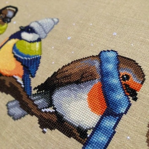 Closeup of Winter Birds cross stitch pattern. Image is zoomed in on the robin. Very cute and very round. The robin is wearing a blue, striped scarf and looks very warm. The robin is facing the wren.