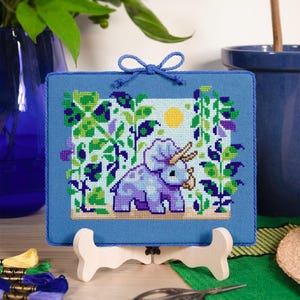 Puede incluir: Un bordado de punto de cruz enmarcado con un triceratops morado en un jard&iacute;n, con un sol amarillo. El marco azul est&aacute; adornado con un lazo azul. La obra de arte se exhibe en un caballete de madera.