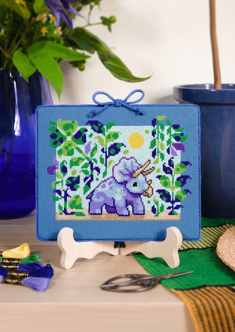 Puede incluir: Un bordado de punto de cruz enmarcado con un triceratops morado en un jard&iacute;n, con un sol amarillo. El marco azul est&aacute; adornado con un lazo azul. La obra de arte se exhibe en un caballete de madera.
