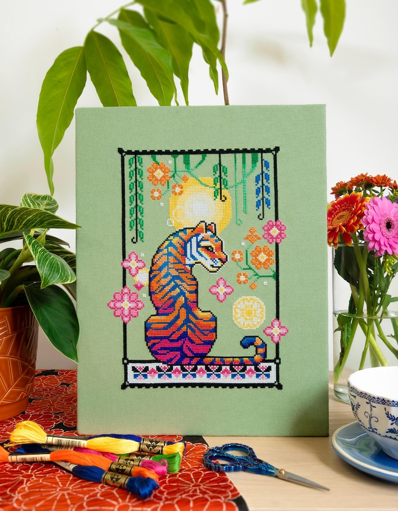 May include: Cross-stitch art of a tiger with blue, orange, and pink stripes, set against a green background. The design includes floral elements and a yellow sun. The artwork is framed in a black border.