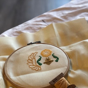 Picture of the work-in-progress of the Egyptian scarab cross stitch pattern. A small part of the pattern has been stitched: only the head, front paws, sun and part of the lefthand wing are visible. Piece is framed in a hoop, bobbin in the foreground.